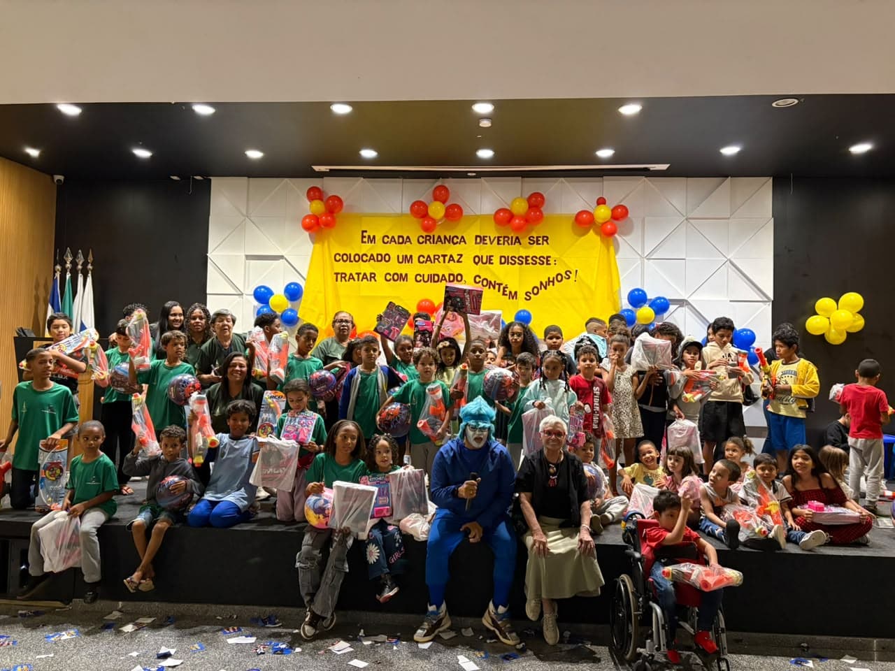 Crianças no palco do auditório segurando os presentes do Dia das Crianças, com o palhaço de azul sentado à frente e balões ao fundo.