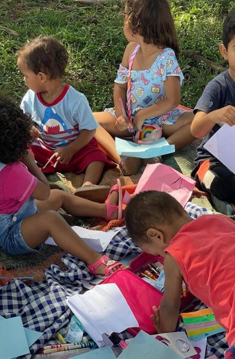 Crianças sentadas na grama, desenhando e pintando em uma atividade ao ar livre.