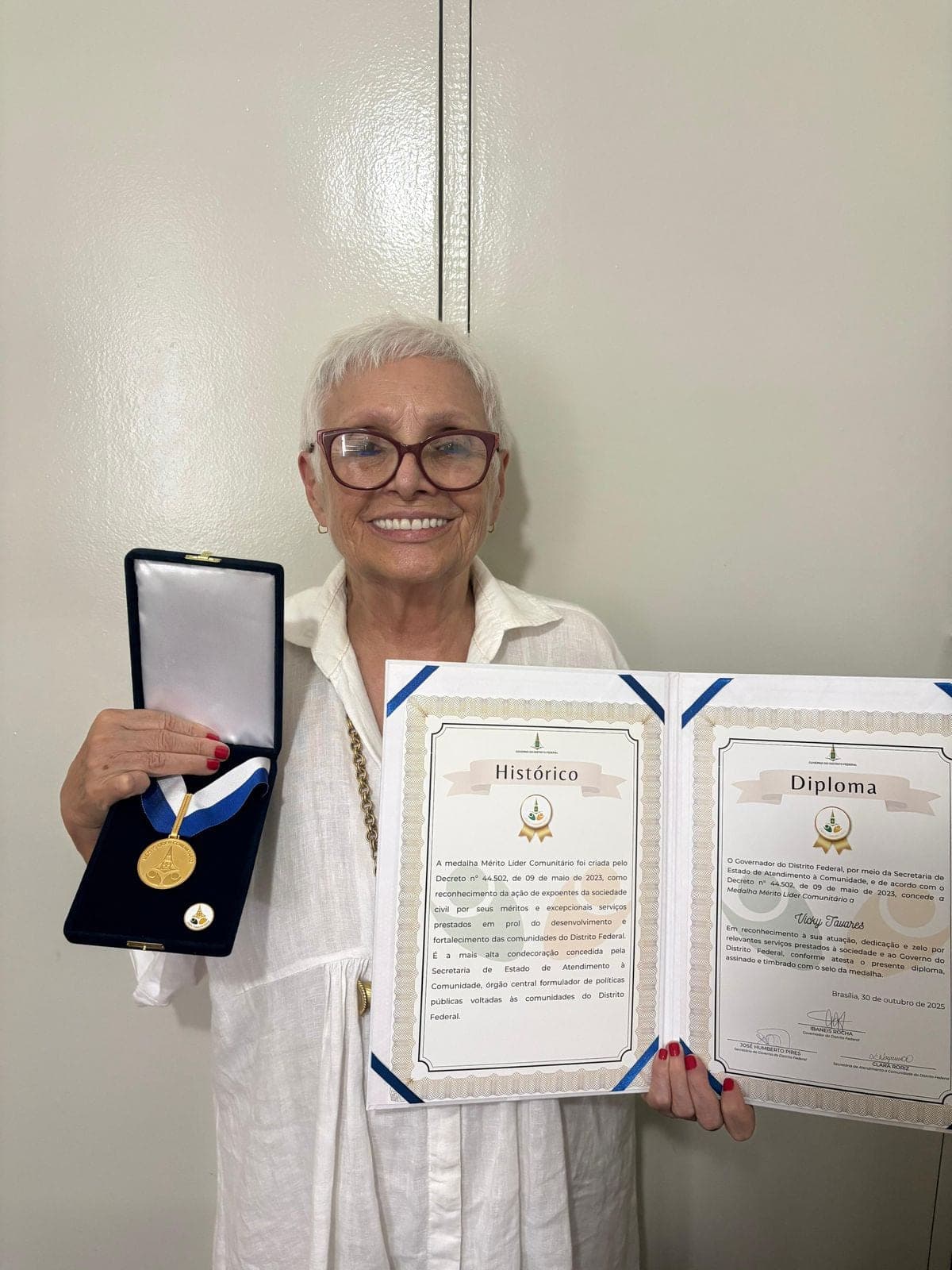 Vicky Tavares sorrindo, segurando a Medalha Mérito Líder Comunitário e o diploma correspondente.